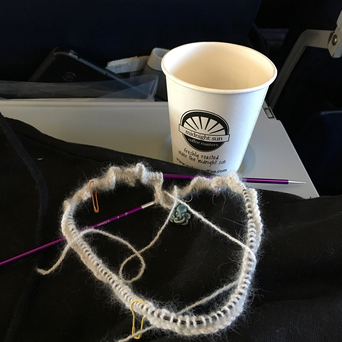 A round of knitting, just cast on, and an empty coffee cup on an airplane seat back table. A tablet sticks out from the seat back pocket in back of the photo.