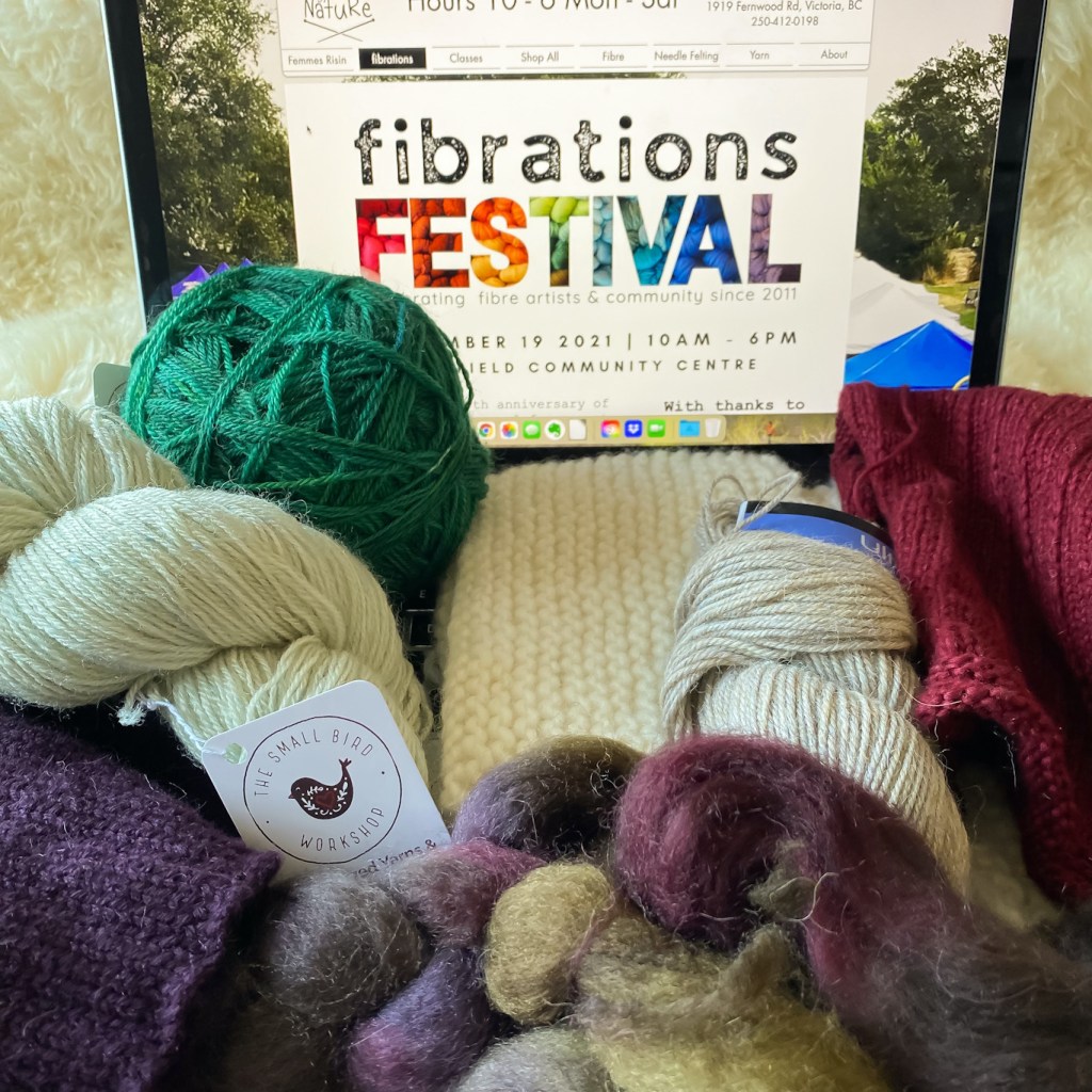 Yarn, spinning fibre, and knitting projects in progress, sitting in front of a computer screen with the splash page of Fibrations Festival showing on it.