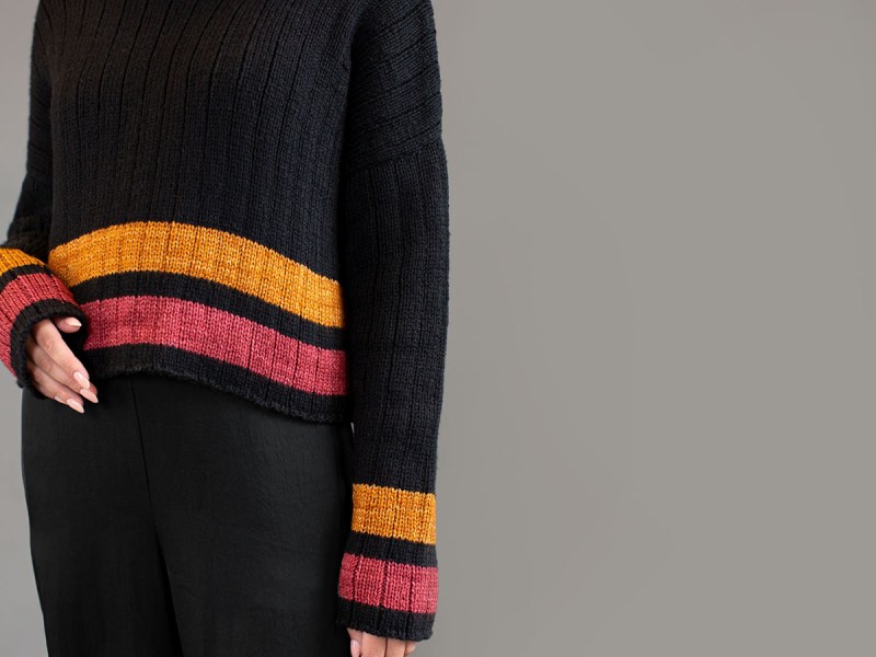 Torso of a woman wearing a black sweater with pink and amber stripes at the cuffs and hem.