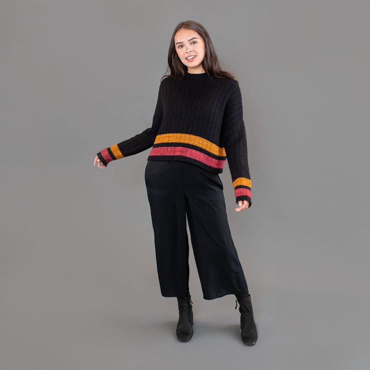 Standing woman wearing a black sweater with pink and amber stripes at the cuffs and hem.