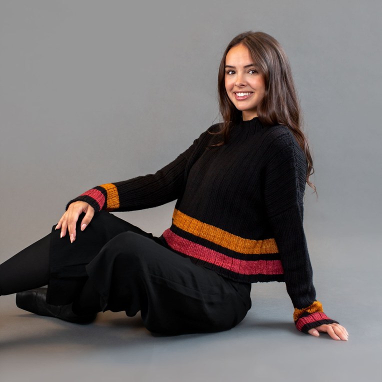 Seated woman wearing a black sweater with amber and pink stripes at the cuffs and hem.