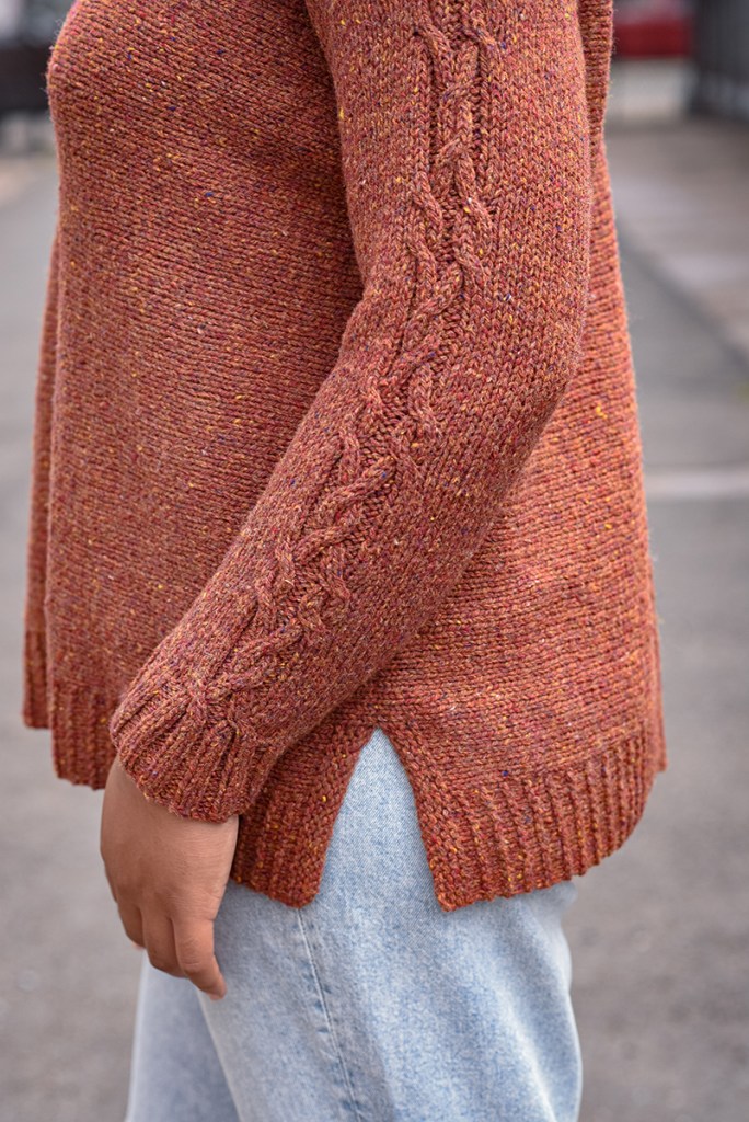 Close up of a woman wearing a hand knit pullover, showing the cable detail on the sleeve and the split hem.