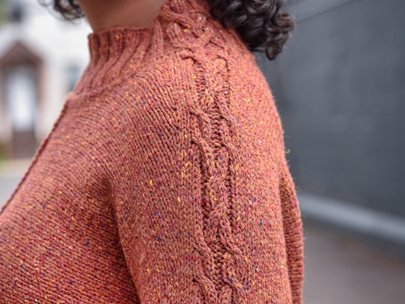 Close up of a woman wearing a hand knit pullover with a cable running up the sleeve into the neckband.