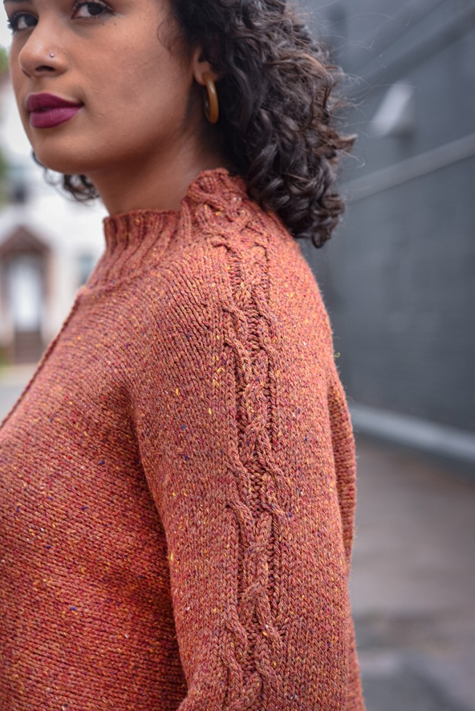 Close up of a woman wearing a hand knit pullover with a cable running up the sleeve into the neckband.