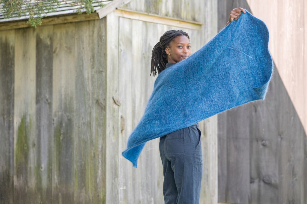 A woman with hands extended, holding out a blue, handknit shawl with a long, wavy line extending the length of the piece.
